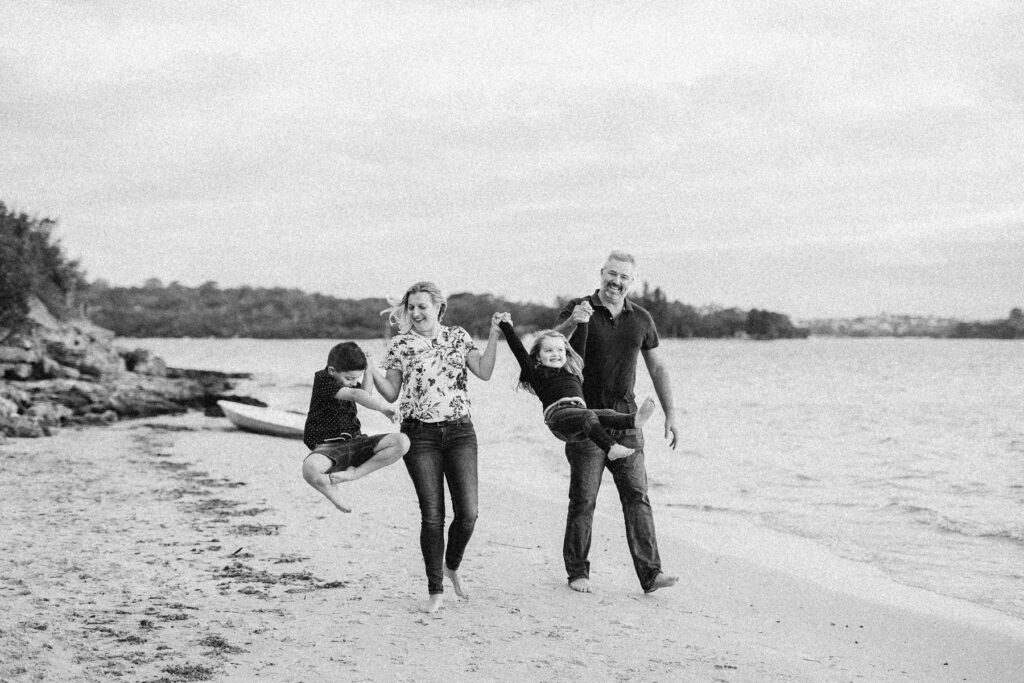 Parents swinging their two children while walking along the beach during a black and white family photography session at Bishop Road Reserve in Perth.