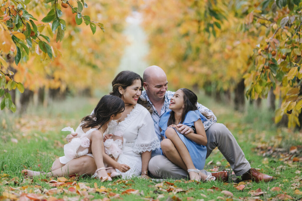 Family sitting together beneath colourful autumn trees during a natural family photography session at a Roleystone orchard near Perth.
